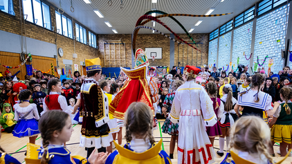 Pänz große Pause 2023 Das Kölner Kinderdreigestirn 2023 ist von hinten zu sehen, von links nach rechts Kinderbauer, Kinderprinz und Kinderjungfrau. Im Vordergrund sieht man die Pagen und Gardisten des Kölner Kinderdreigestirns in blau-gelben Uniformen. Im Hintergrund sind unzählige Kinder zu sehen, die tanzen und singen und jubeln. Das Foto wurde in einer Grundschulturnhalle aufgenommen.