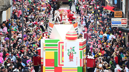 Das Kölner Kinderdreigestirn im Kölner Rosenmontagszug auf der Severinstraße Man sieht von oben auf die Severinstraße direkt hinter der Severinstorburg herab. Rechts und links von der Straße stehen mehrere Reihen bunte Zuschauer, auf der Straße ist der Wagen des Kölner Kinderdreigestirns. Der Wagen ist bunt und mit dem Jecko geschmückt, man kann das Kölner Kinderdreigestirn sehen.